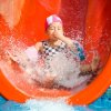 Bambino scivola divertito su uno scivolo d'acqua rosso, creando spruzzi in piscina.