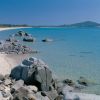 Spiaggia con sabbia chiara, mare blu e grandi rocce sul bagnasciuga.