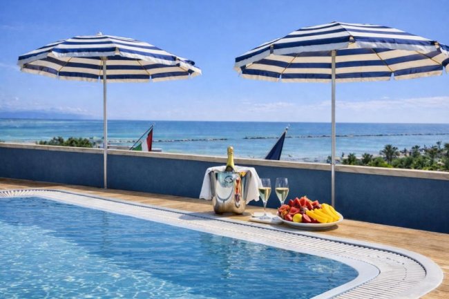 Piscina con ombrelloni a strisce, vista mare, e tavolino con frutta e vino.