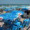 Ombrelloni e sedie blu su una terrazza sul mare con la spiaggia affollata sullo sfondo.