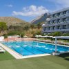 Piscina con lettini e ombrelloni, un hotel sullo sfondo e montagne in lontananza.