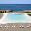 Una grande piscina vista dall'alto con lettini e ombrelloni bianchi e mare sullo sfondo.